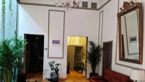 Un salón amplio con un sofá rojo, una silla negra y un espejo grande. Hay varias plantas en macetas y una araña.