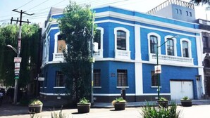 Een blauw gebouw met witte accenten, met een centrale ingang en balkon, omringd door potplanten.