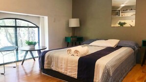 Een net opgemaakt bed met een opgerolde handdoek, een nachtkastje met een lamp, een kleine stoel en een groot raam met uitzicht op bomen.