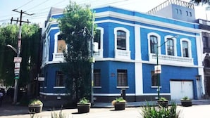 A blue building with white trim, featuring a central entrance with a balcony, surrounded by potted plants.