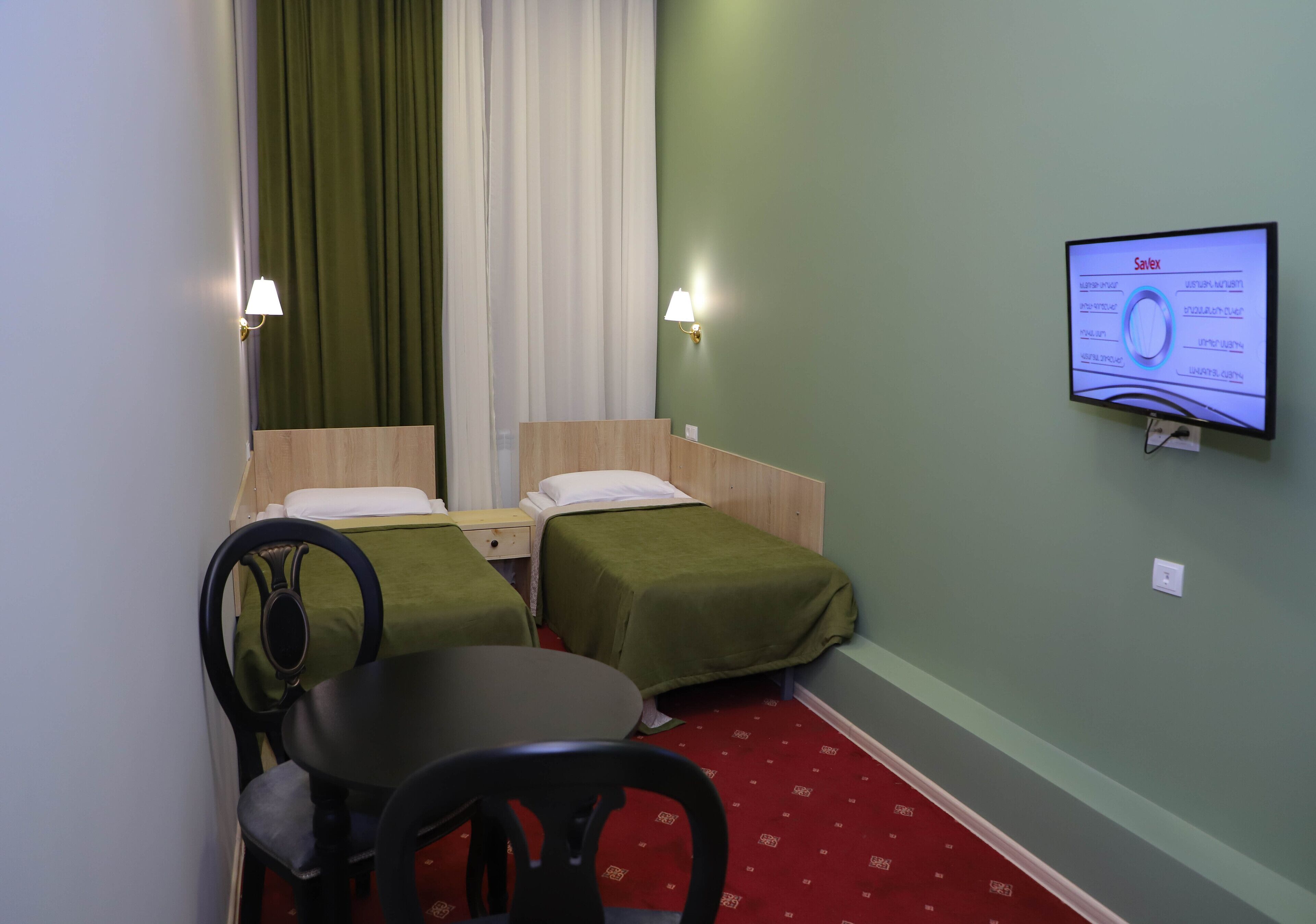 Habitación de hotel con dos camas individuales, un escritorio con silla, un televisor en la pared y una alfombra roja.