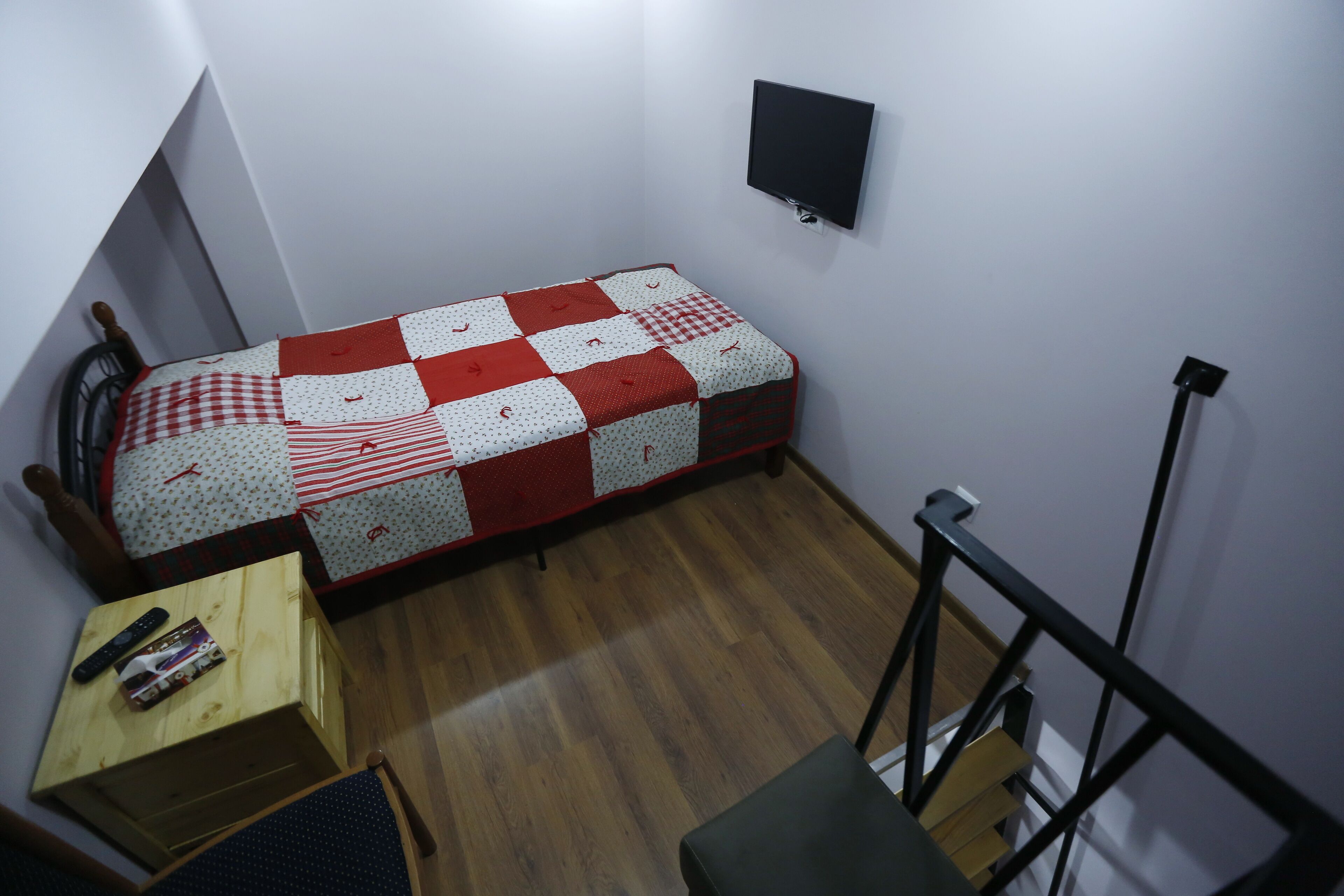 A small, compact room with a wooden floor, a bed with a red and white checkered quilt, a small wooden table with a remote control, and a black mounted TV on the wall.