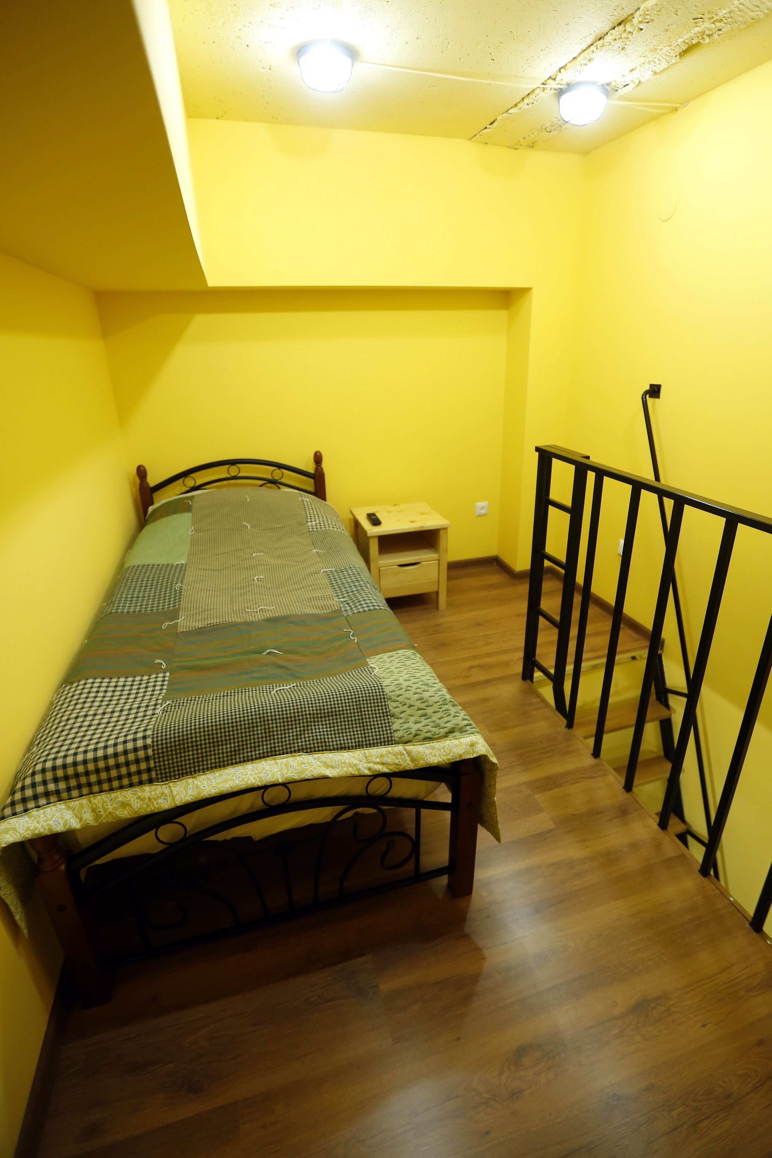 A small, narrow room with a single bed, a wooden nightstand, and a black metal railing.