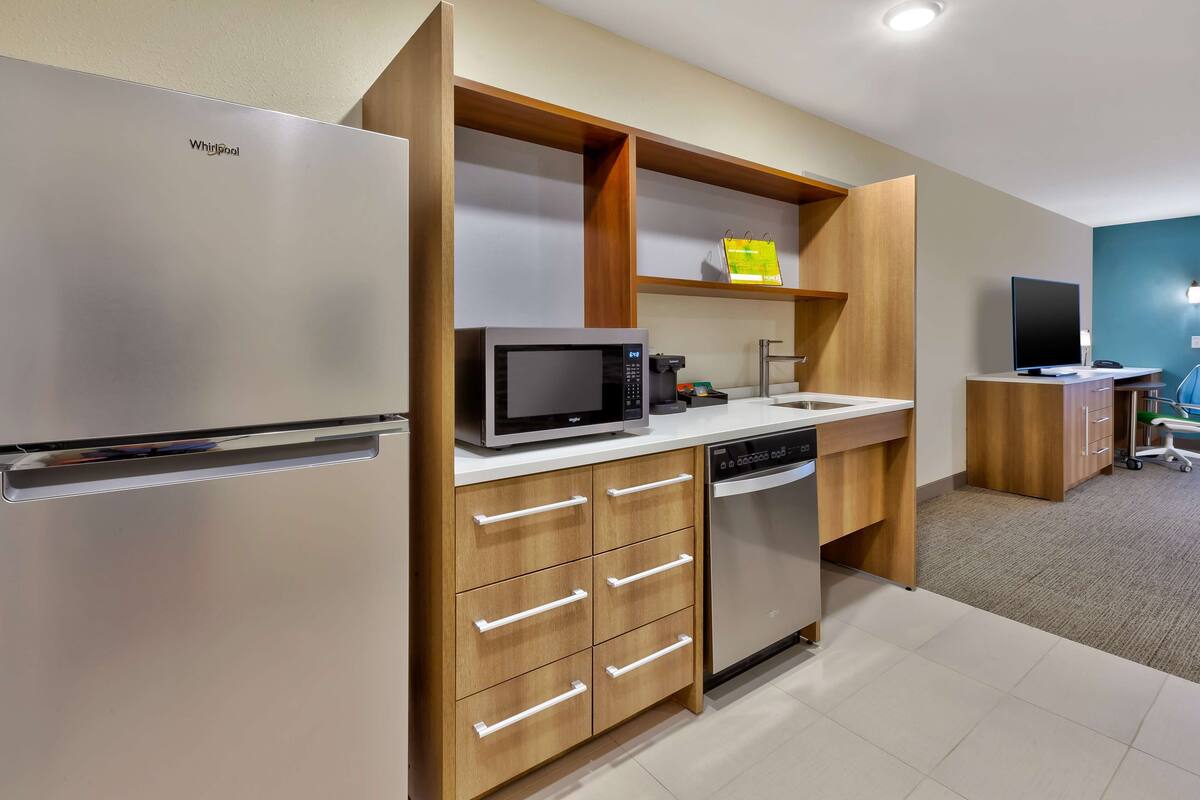 Eine moderne Küche mit einem Edelstahlkühlschrank, einer Mikrowelle, einem Geschirrspüler, Holzschränken und einem Holzregal mit einem gelben Gegenstand darauf.