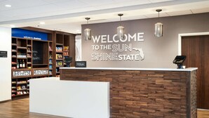 A reception desk with a "Welcome to the Sun Shine State" sign, a Hilton Honors logo, and a variety of amenities including a mini bar and a minibar. - Hampton Inn Clewiston (Clewiston)