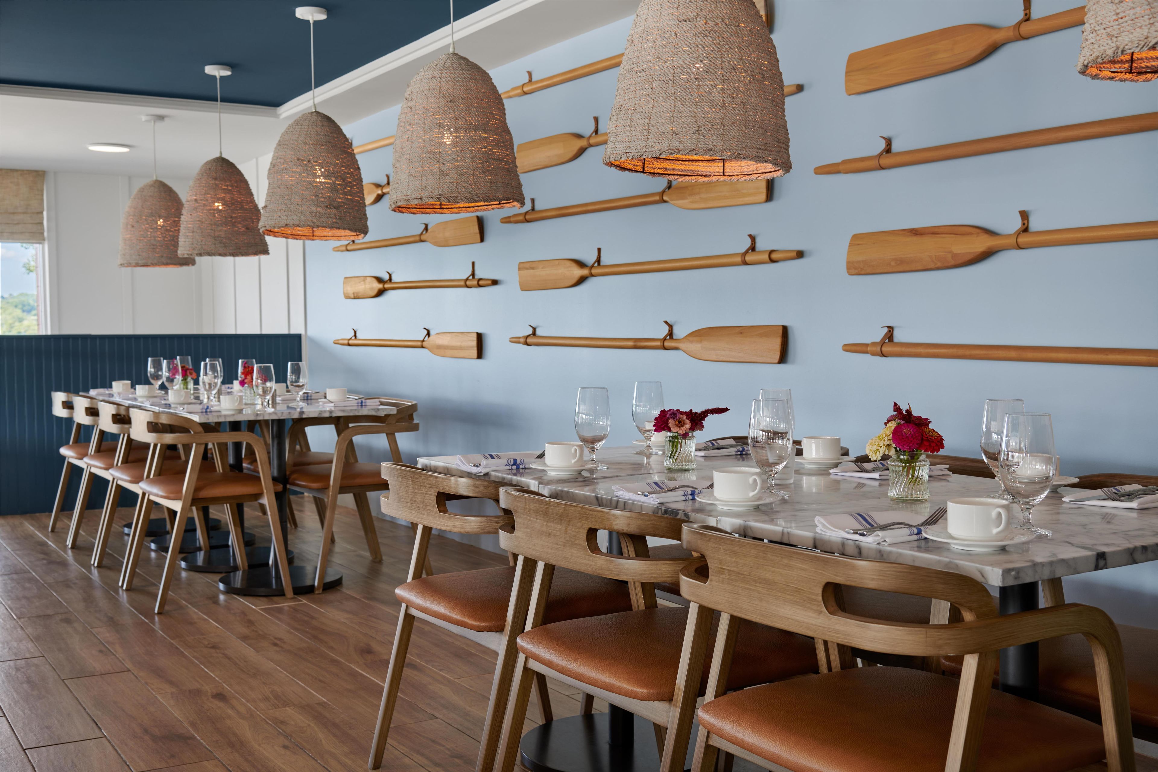 A dining area with wooden tables and chairs, a wall-mounted wooden paddle display, and hanging woven light fixtures.