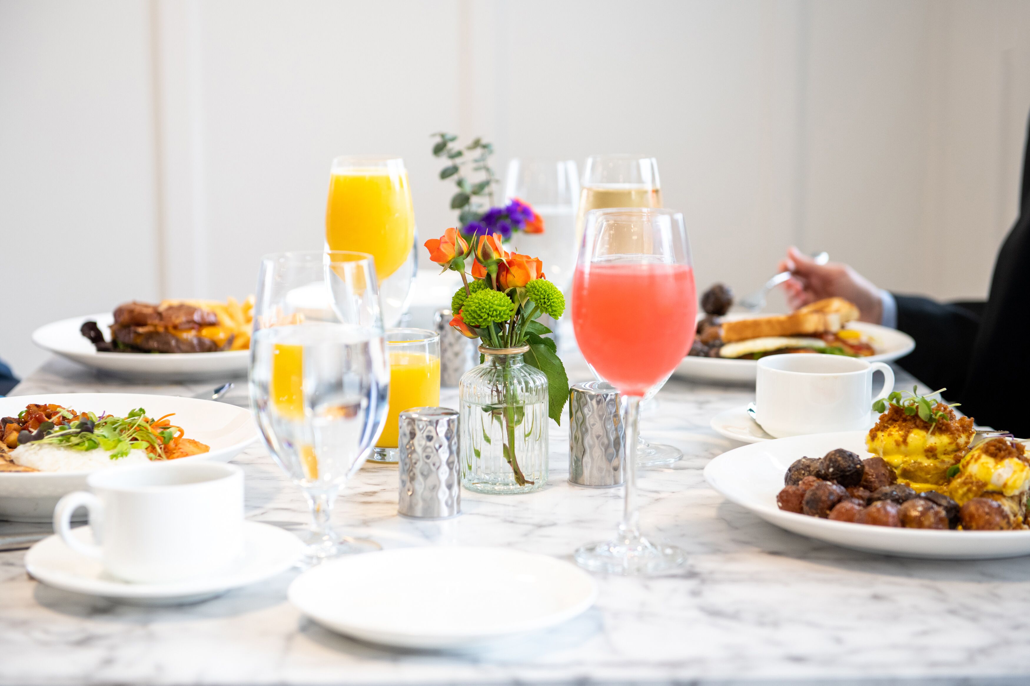 A breakfast setting with various dishes, drinks, and a vase of flowers on a marble table.