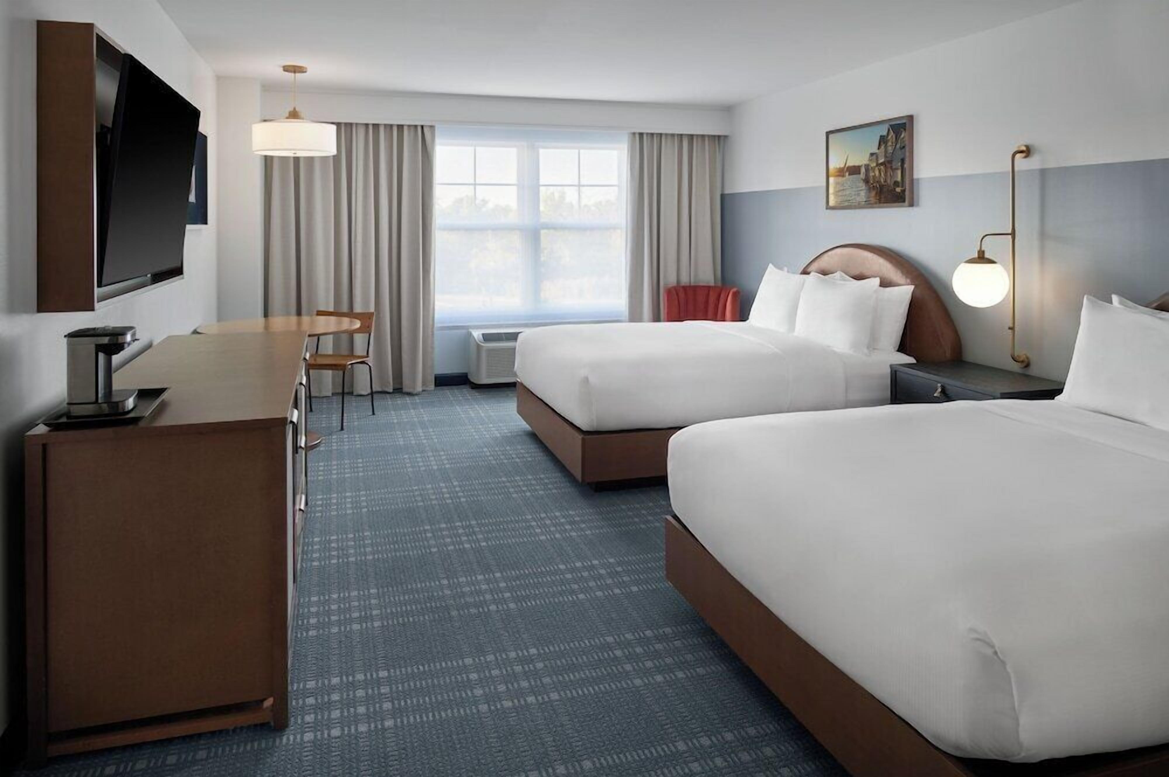 A hotel room with two beds, a desk, a TV, a window with curtains, and a painting on the wall.