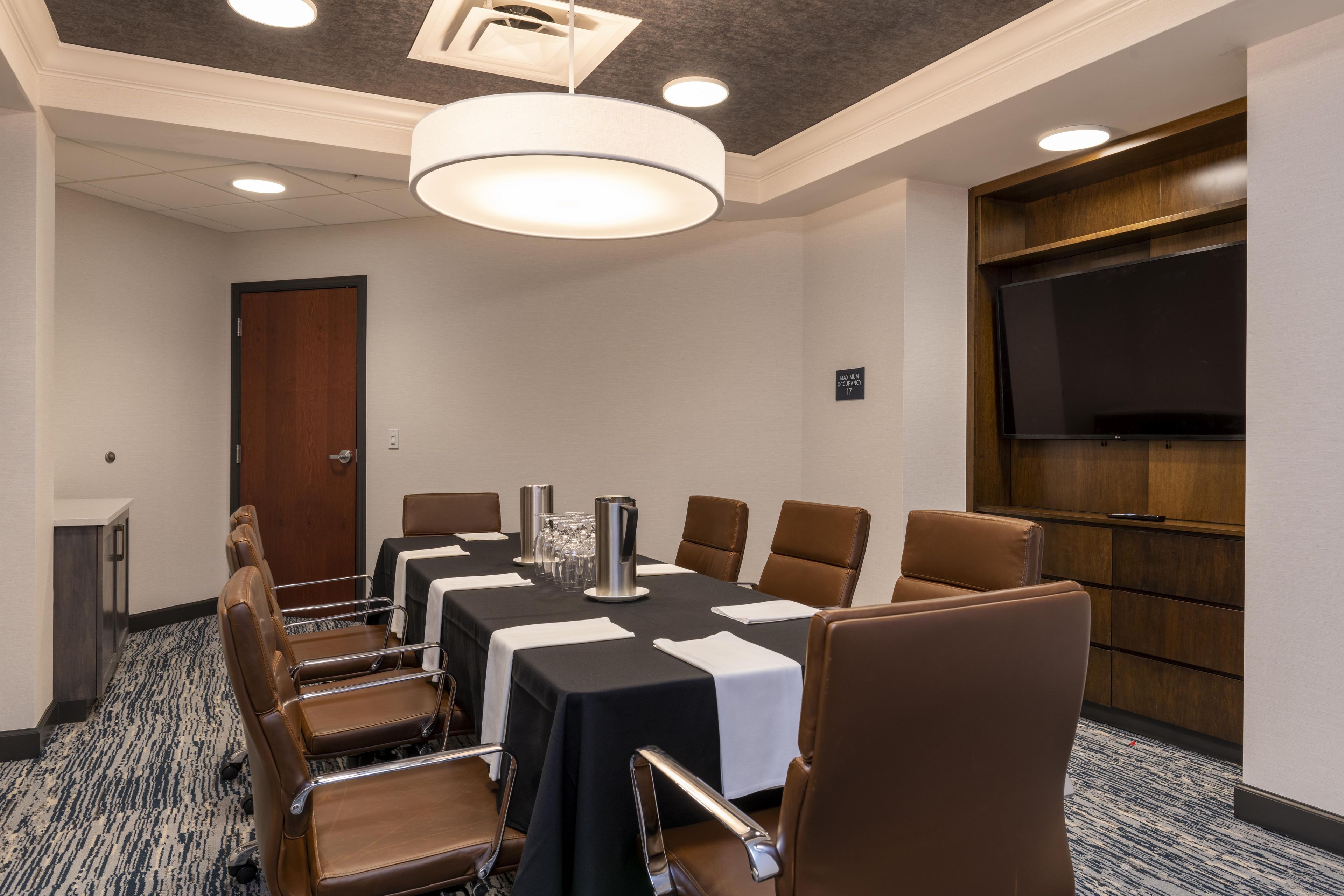 A conference room with a long table, brown leather chairs, and a television on the wall.