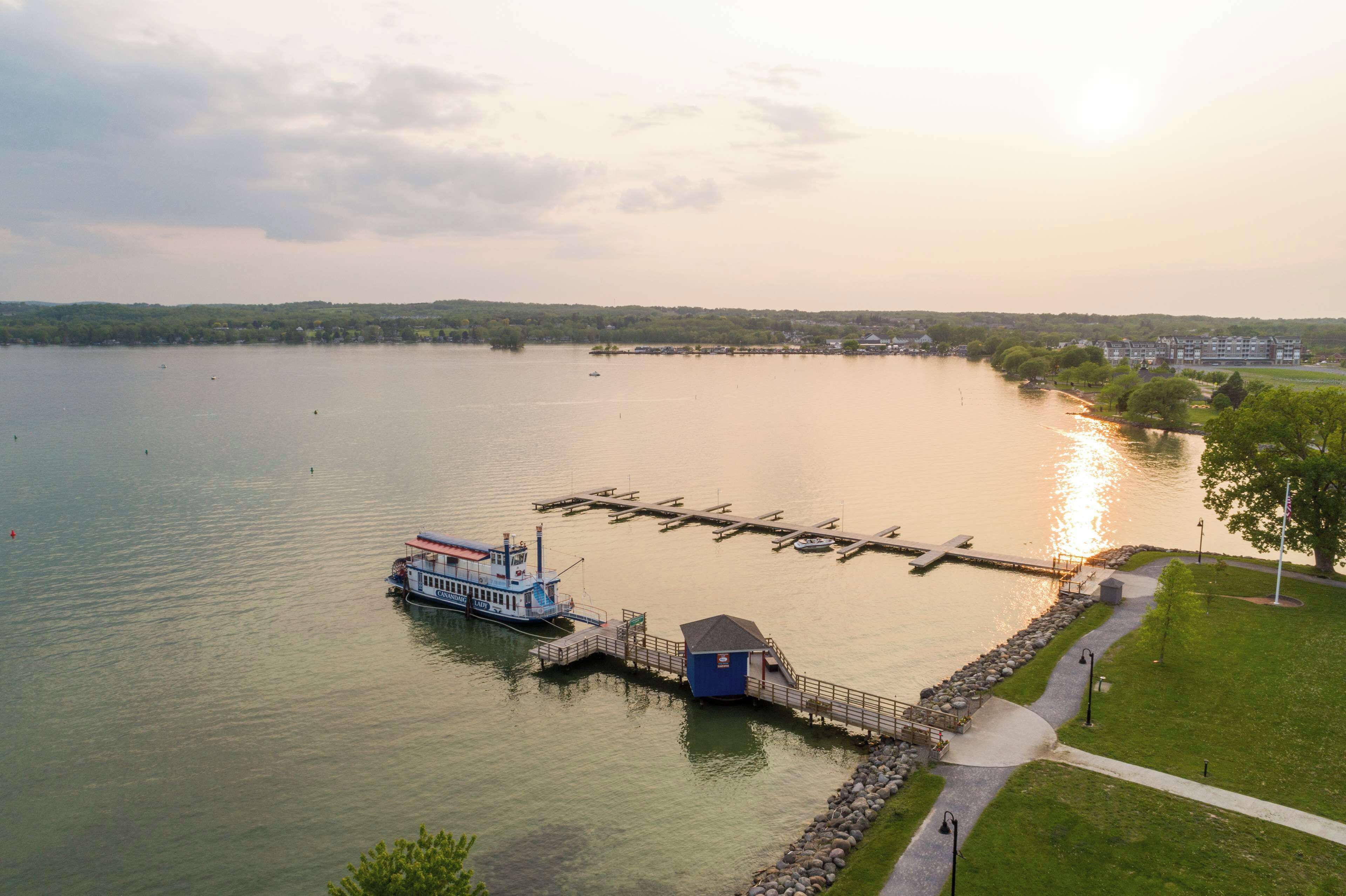 A boat docked at a pier with a small building nearby, surrounded by calm water and a grassy area with a path.