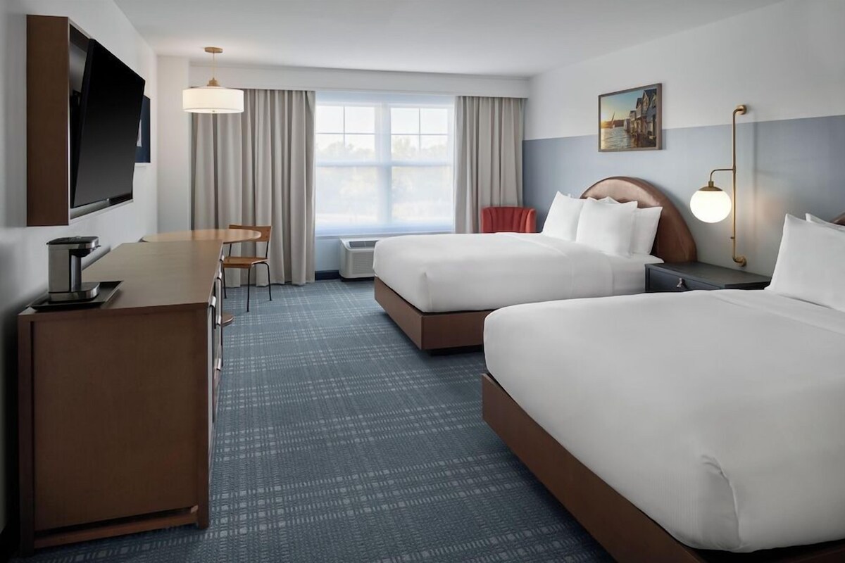 A hotel room with two beds, a desk, a TV, a window with curtains, and a painting on the wall.