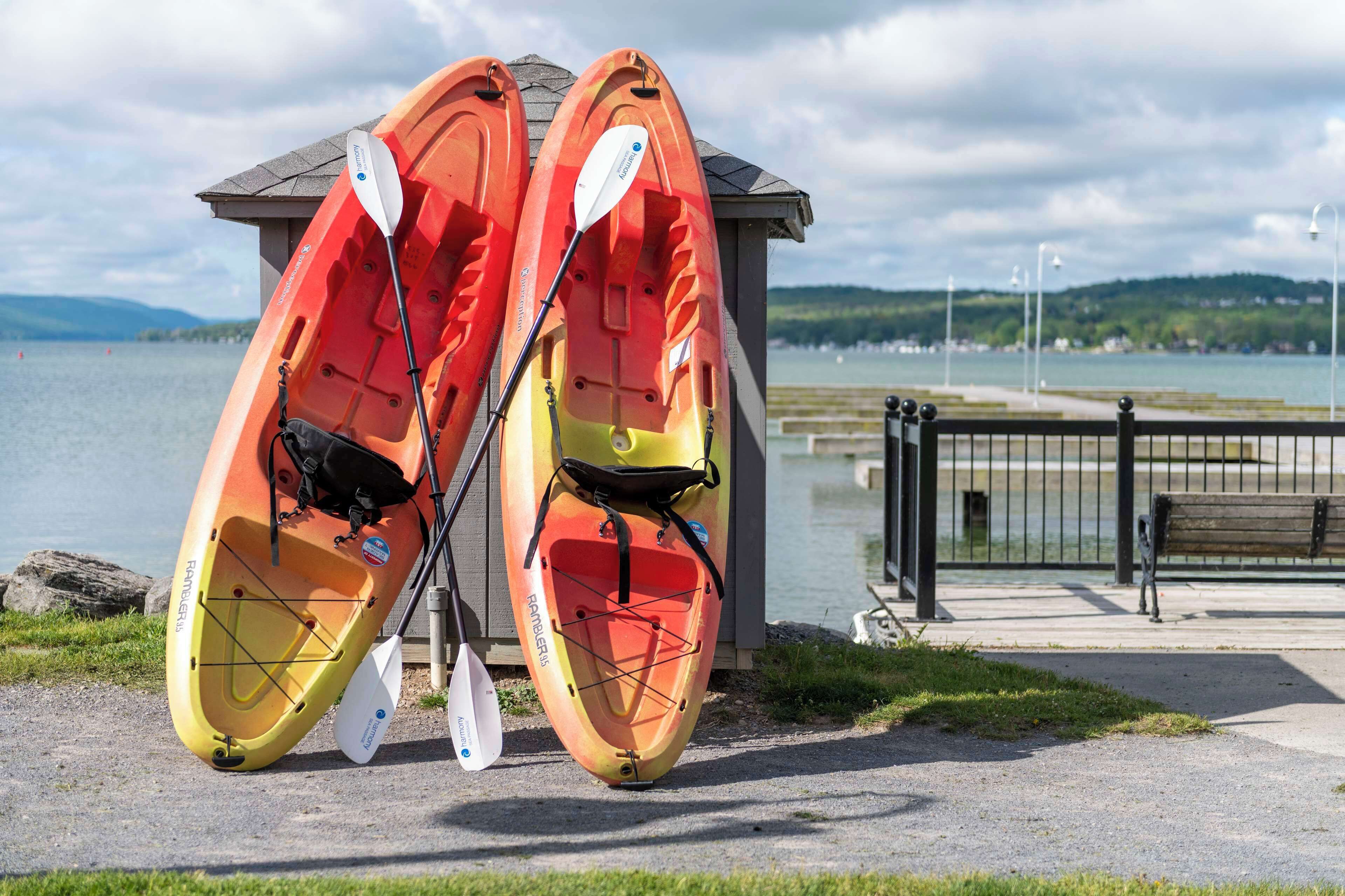 Two kayaks with paddles leaning against a small shed by a lakeside walkway.