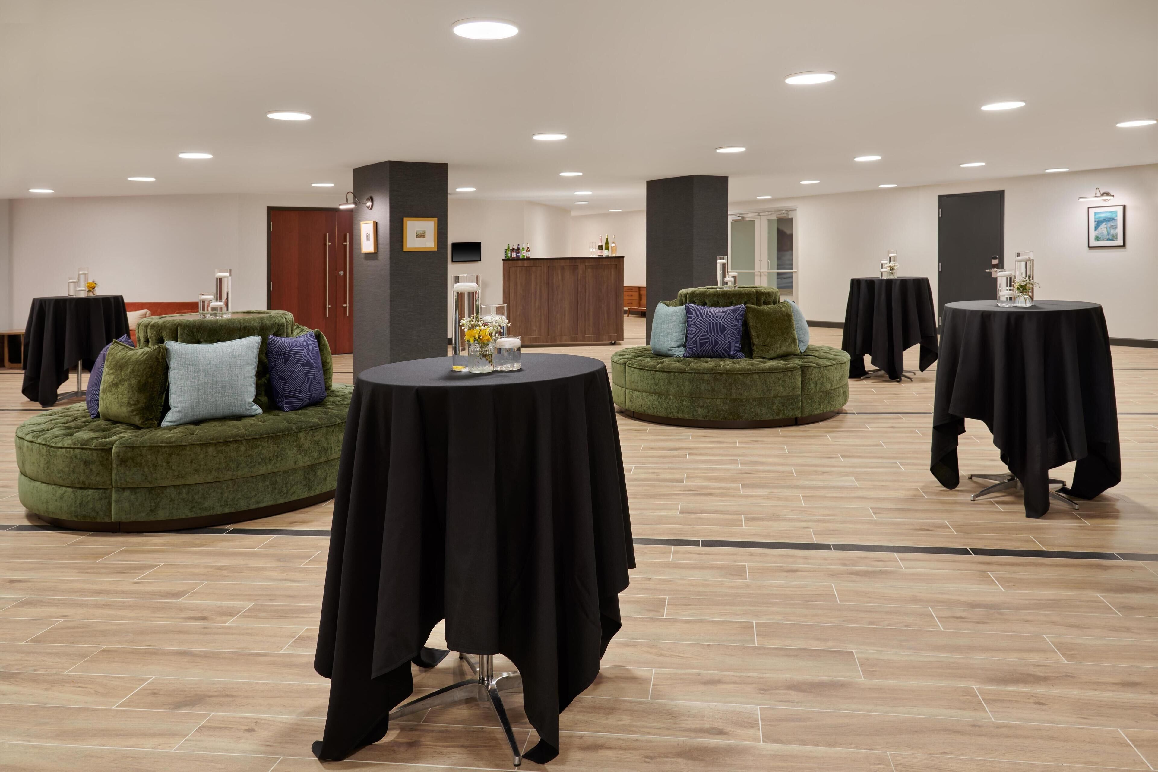 A spacious room with multiple round tables covered with black tablecloths, green upholstered sofas with cushions, and a wooden floor.