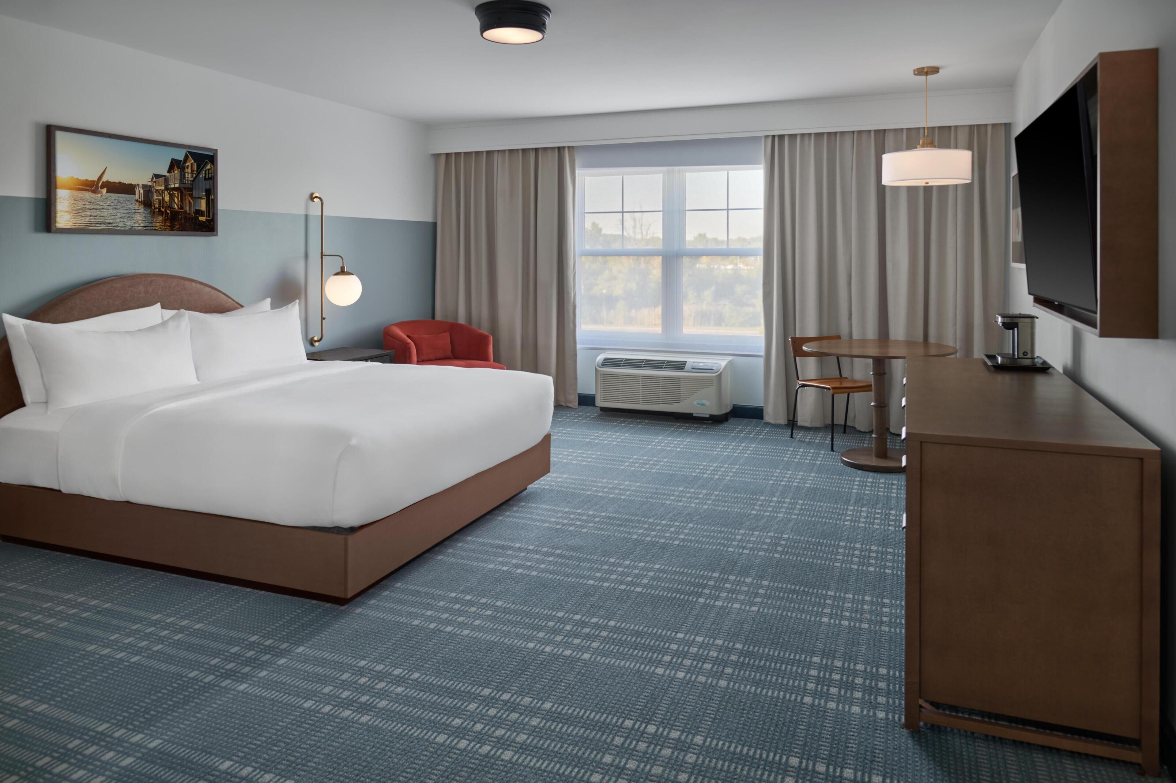 Habitación de hotel con una cama grande, un escritorio con una silla, un televisor y un ventanal con cortinas.