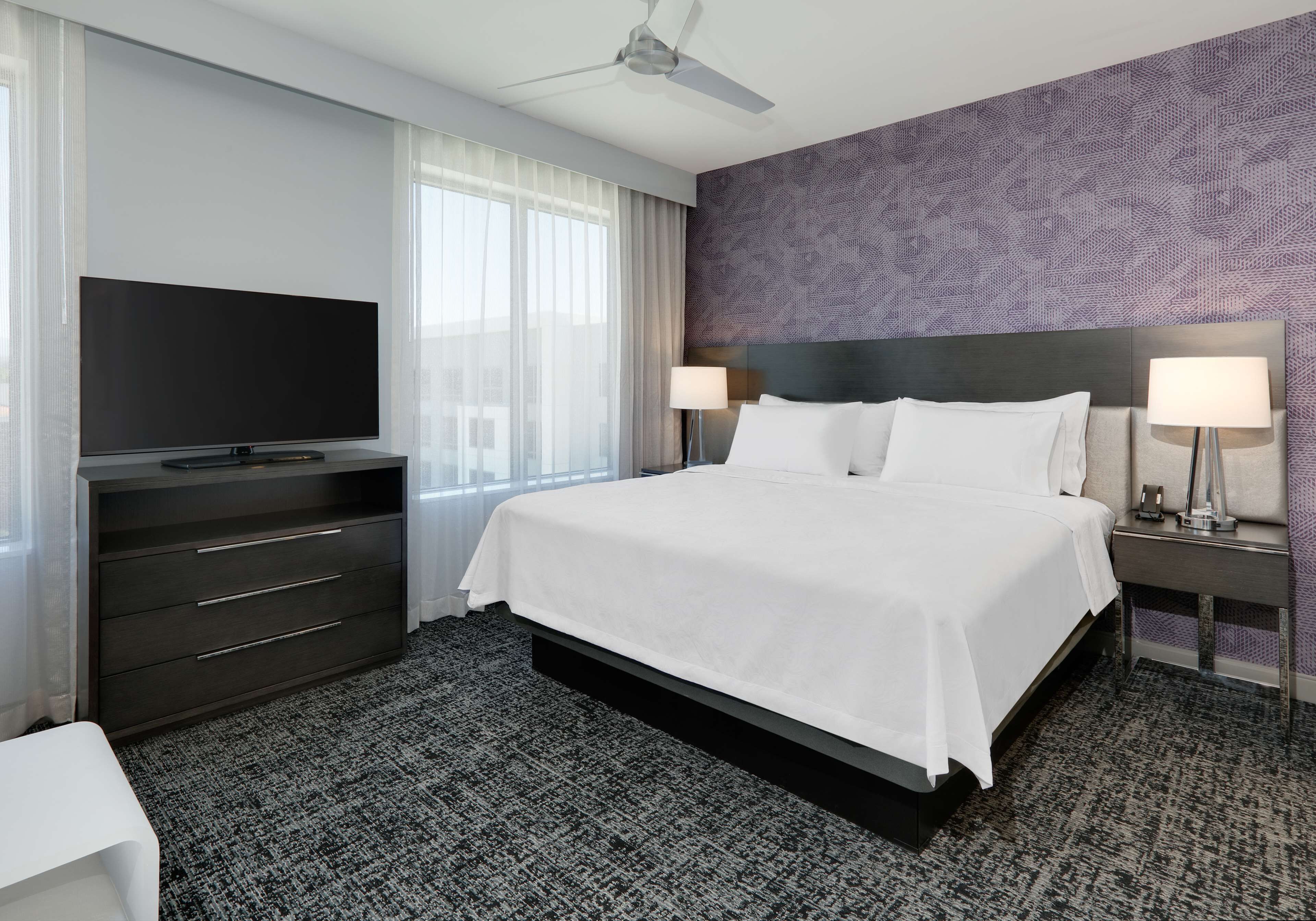 Habitación de hotel con una cama grande, un televisor de pantalla plana, una cómoda, dos lámparas de noche y un ventilador de techo.