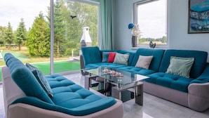 Una sala de estar moderna con un sofá color turquesa, una mesa de centro de vidrio y vistas a un jardín a través de amplios ventanales.