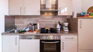 Une cuisine moderne équipée d’une cuisinière, d’un four et d’un lave-vaisselle en acier inoxydable. On y trouve des armoires en bois, un plan de travail en bois, ainsi que divers ustensiles de cuisine, comme une bouilloire, une machine à café et des légumes.