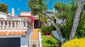 A white building with a tiled roof, a covered porch, and a small garden with a palm tree and yellow shrubs. - Beautiful  villa for 6 guests with private pool, WIFI, A/C, TV and patio (Calafell)