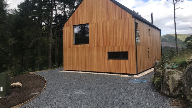 Una cabaña de madera con techo inclinado, camino de grava y un bosque al fondo.