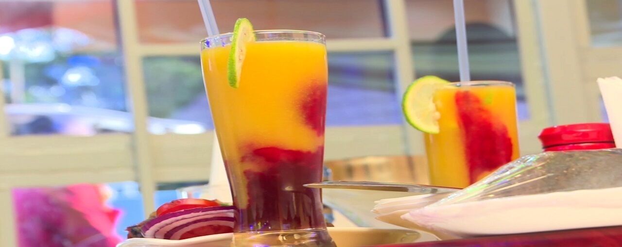 Two tall glasses of fruit juice with lime garnishes, placed on a table with a window view in the background.