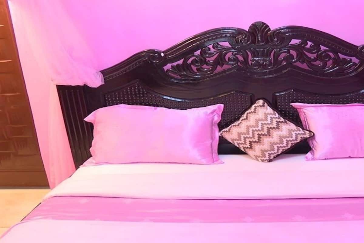 Una cama con cabecera oscura, ropa de cama rosa y un cojín estampado.