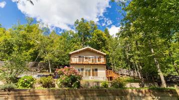 Une cabane en bois à deux étages, dotée d’un balcon, et entourée d’une végétation luxuriante et d’arbres.