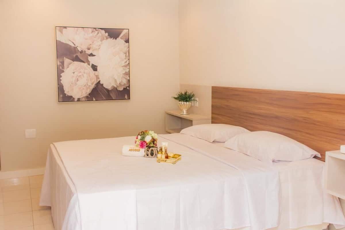 A neatly made bed with white linens, a small bedside table with a plant, and a framed floral artwork on the wall.