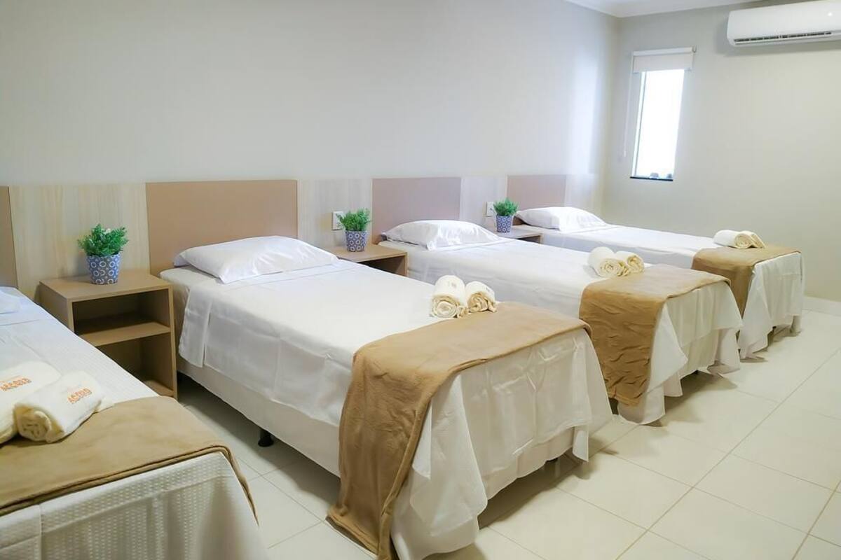 A hotel room with four single beds, each with a white bedspread and a beige blanket. There are small potted plants on the nightstands, and an air conditioning unit on the wall.