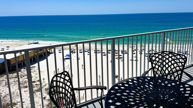Een balkon met metalen stoelen, uitkijkend op een strand met parasols en mensen.