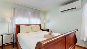 A bedroom with a wooden bed, white bedding, two bedside lamps, a window with sheer curtains, and an air conditioning unit on the wall. - Hank's Hideaway by Bayley Vacation Rentals (Scarborough)