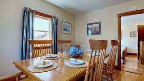 A dining area with a wooden table set for four, surrounded by wooden chairs. There are framed pictures on the walls, a window with curtains, and a door leading to another room.