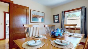 A dining room with a wooden table set for four, surrounded by chairs. There are framed pictures on the wall, a window with curtains, and a view of the outdoors. - Hank's Hideaway by Bayley Vacation Rentals (Scarborough)