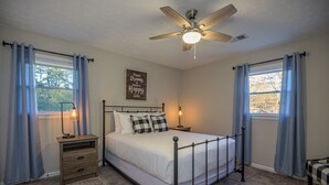 Ein Schlafzimmer mit einem Bett, einem Nachttisch, einem Deckenventilator und zwei mit Vorhängen versehenen Fenstern.