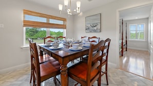 Une salle à manger avec une table en bois dressée pour six personnes, un lustre et une fenêtre avec des stores.