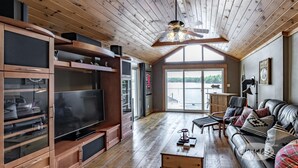 Ein geräumiges Wohnzimmer mit einem großen Flachbildfernseher, Holzböden, einem Ledersofa und einem großen Fenster mit Blick ins Freie.