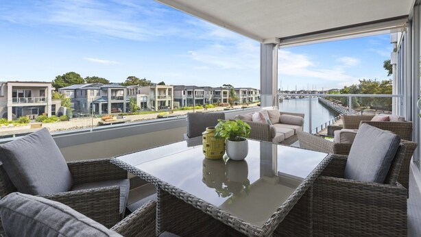 A balcony with wicker furniture, a glass table, and a view of modern buildings and a marina.
