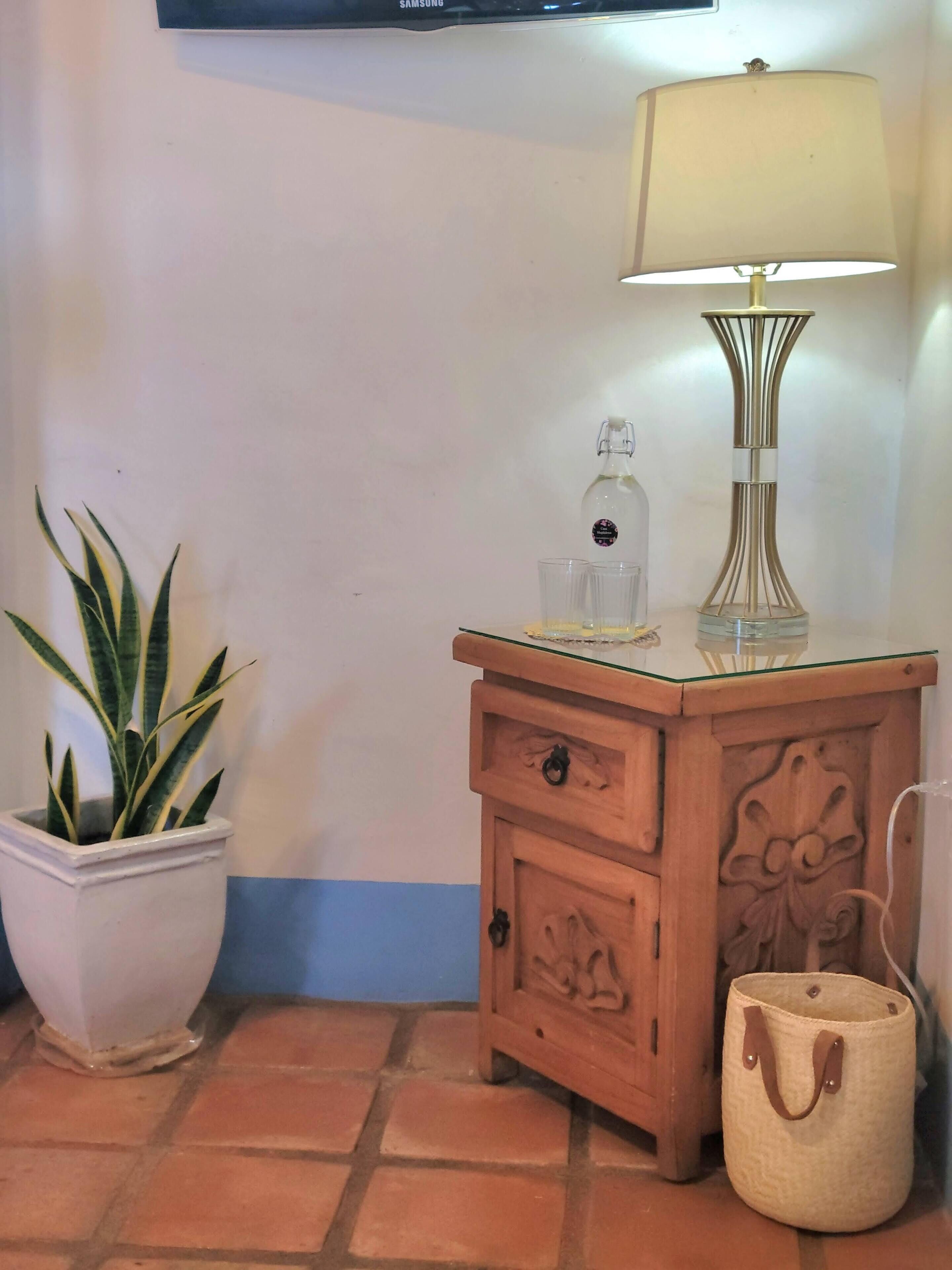 A corner of a room with a wooden nightstand, a lamp, a potted plant, and a small bag.