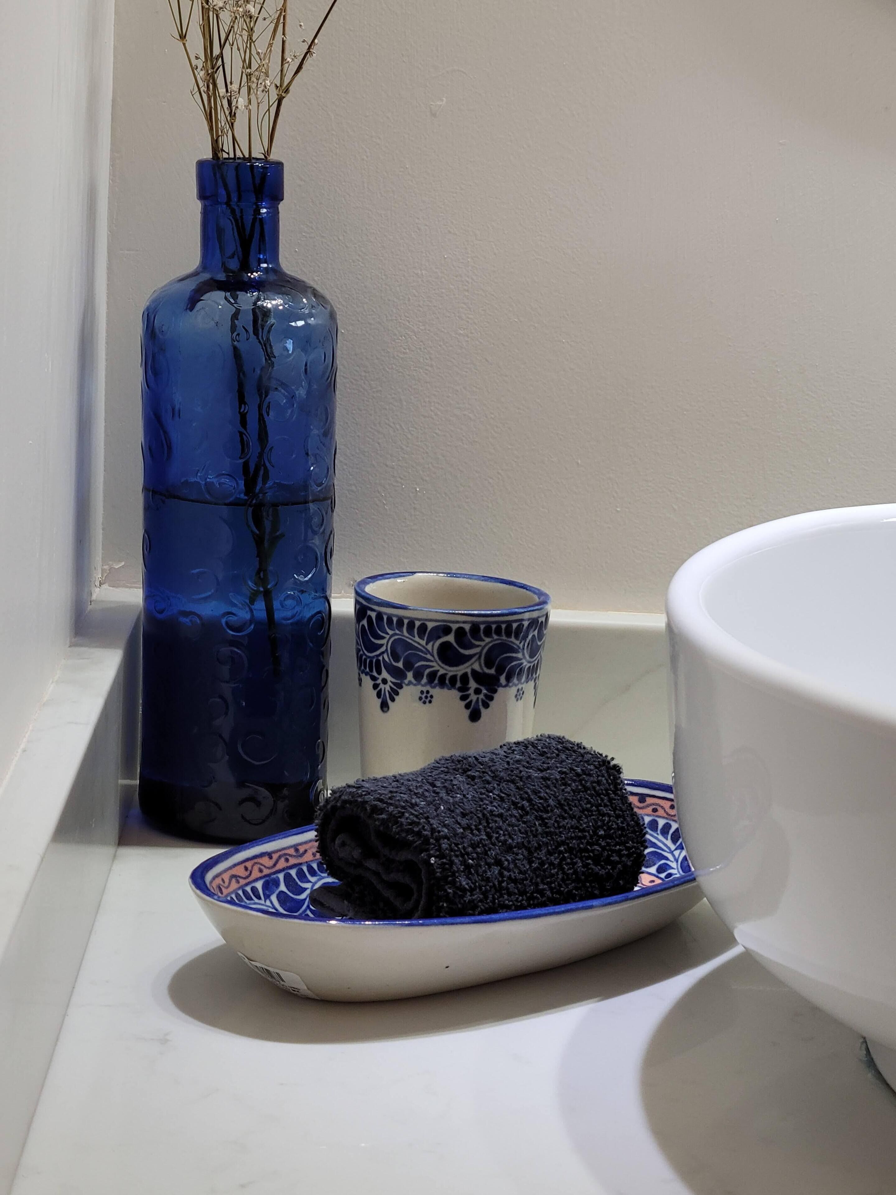 Un baño con un lavamanos blanco, un jarrón azul con plantas secas, una taza decorativa azul y blanca, y una toalla enrollada sobre una bandeja blanca.
