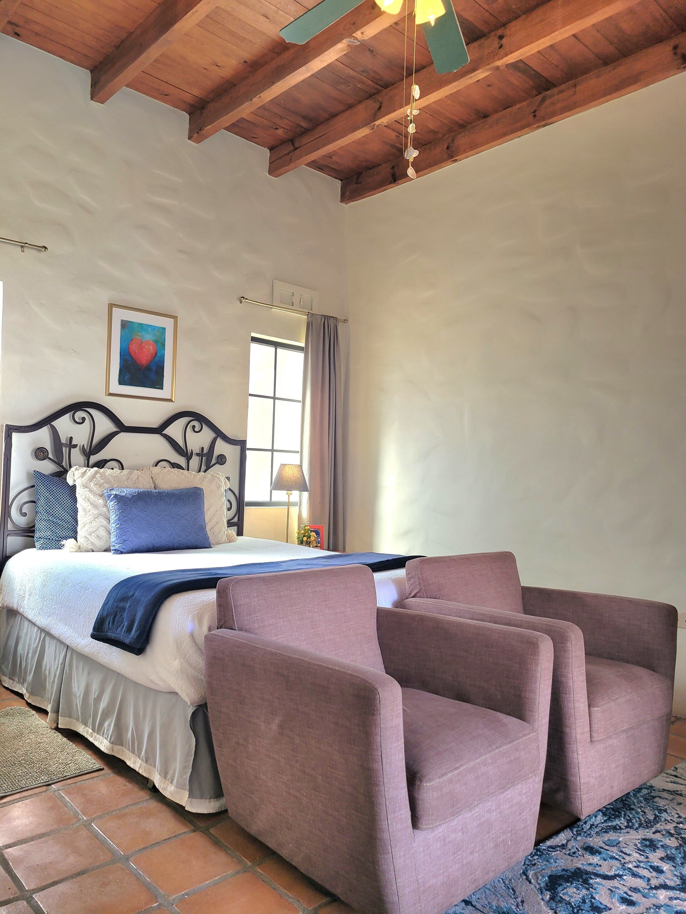 Une chambre à coucher avec un lit, une tête de lit, une fenêtre avec des rideaux, une lampe de chevet, un tableau encadré au mur et un tapis à motifs.