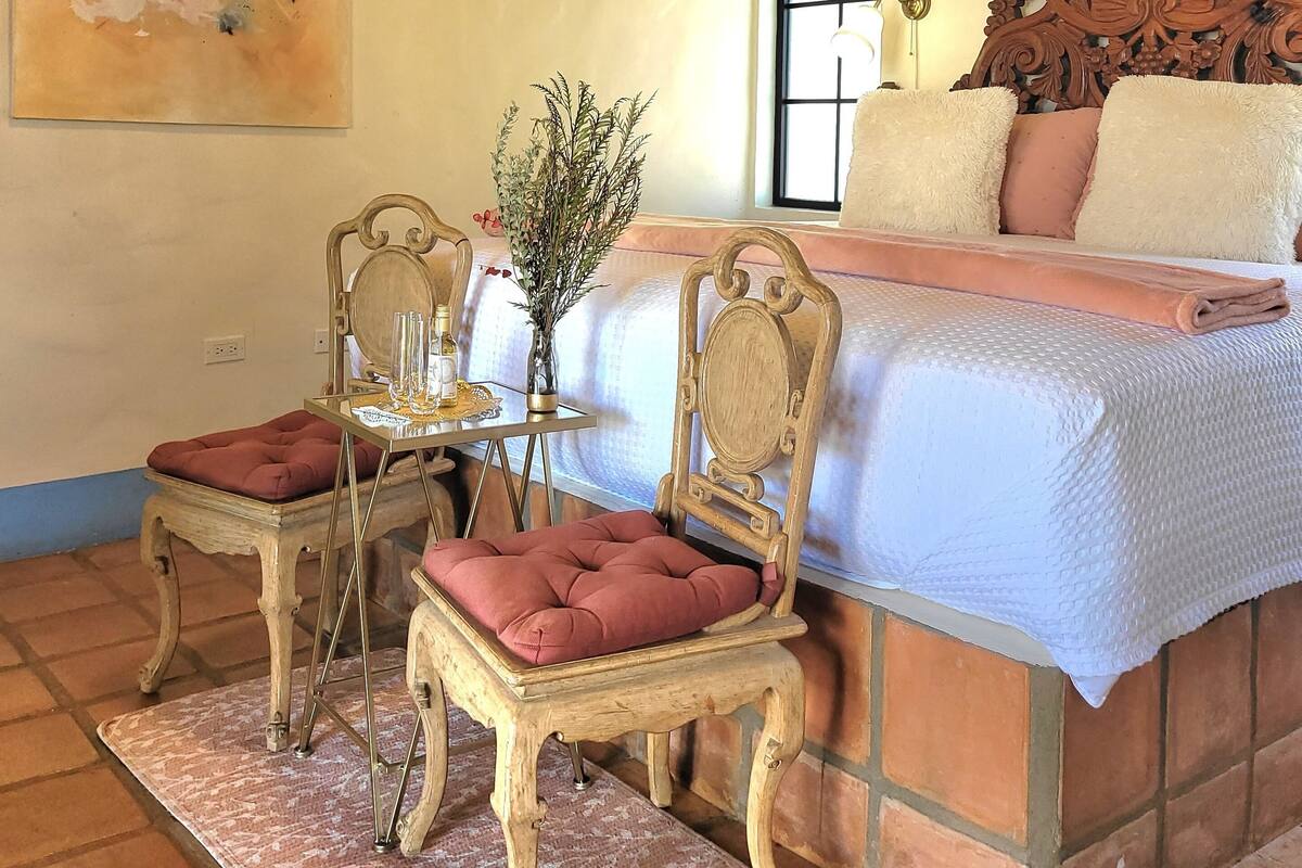 Ein Schlafzimmer mit einem großen Bett, zwei Stühlen, einem kleinen Tisch mit einer Vase und einem Gemälde an der Wand.