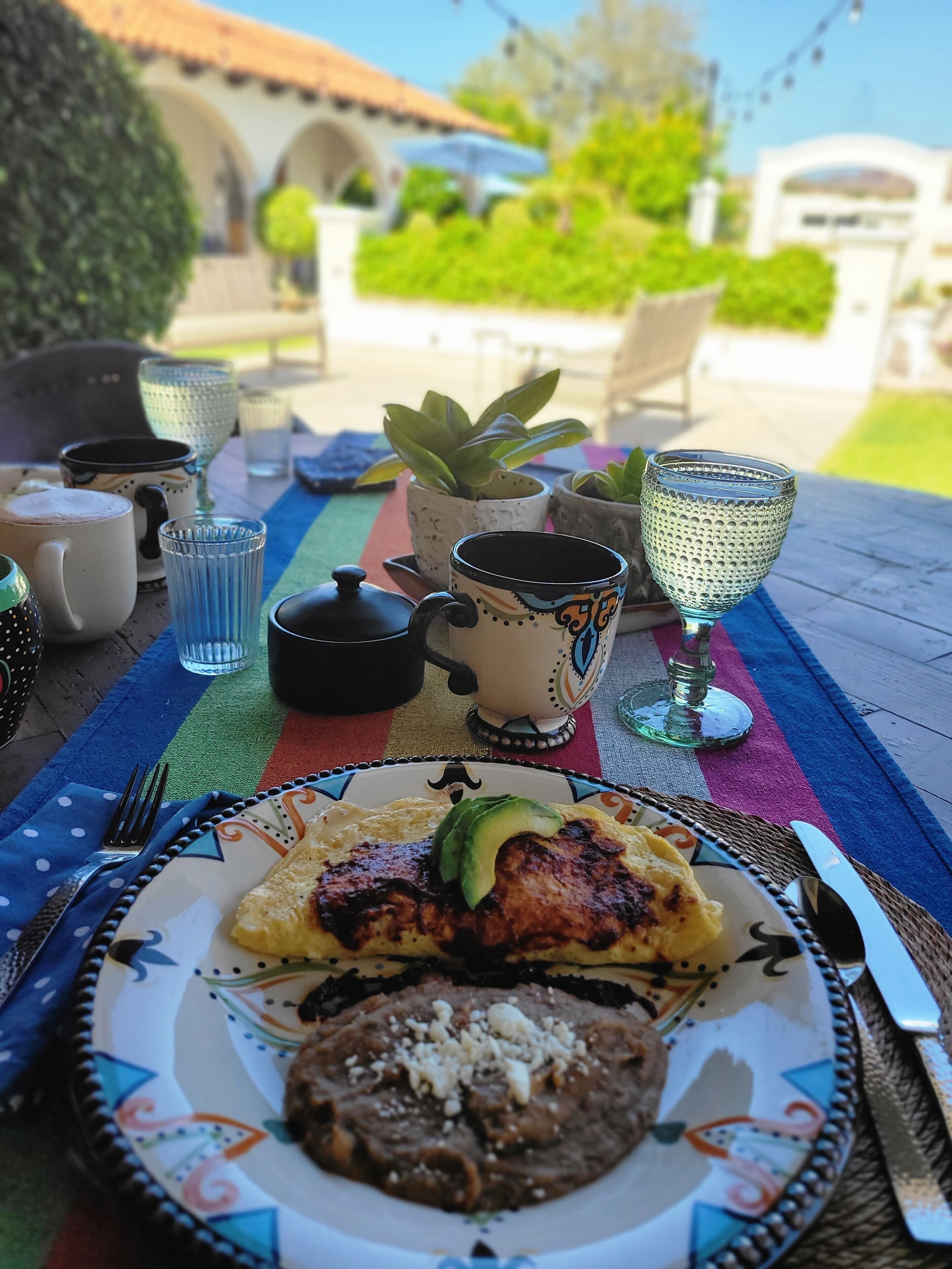 Una mesa dispuesta con un plato de comida, que incluye una hamburguesa a la parrilla y aguacate, rodeada de varias tazas y una tetera.