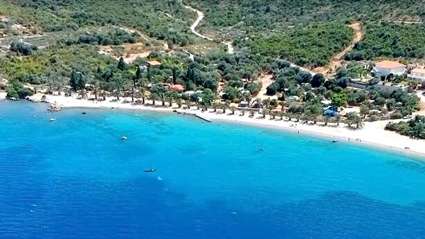 Vista aérea de una zona costera con playa, aguas azules cristalinas y edificios dispersos.