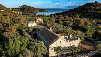 Un bâtiment en pierre, doté d’un toit en tuiles, est entouré d’arbres denses et on aperçoit une étendue d’eau au loin.