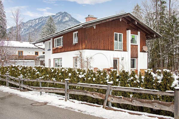 A two-story wooden house with a white facade, a balcony, and a view of snow-covered mountains.