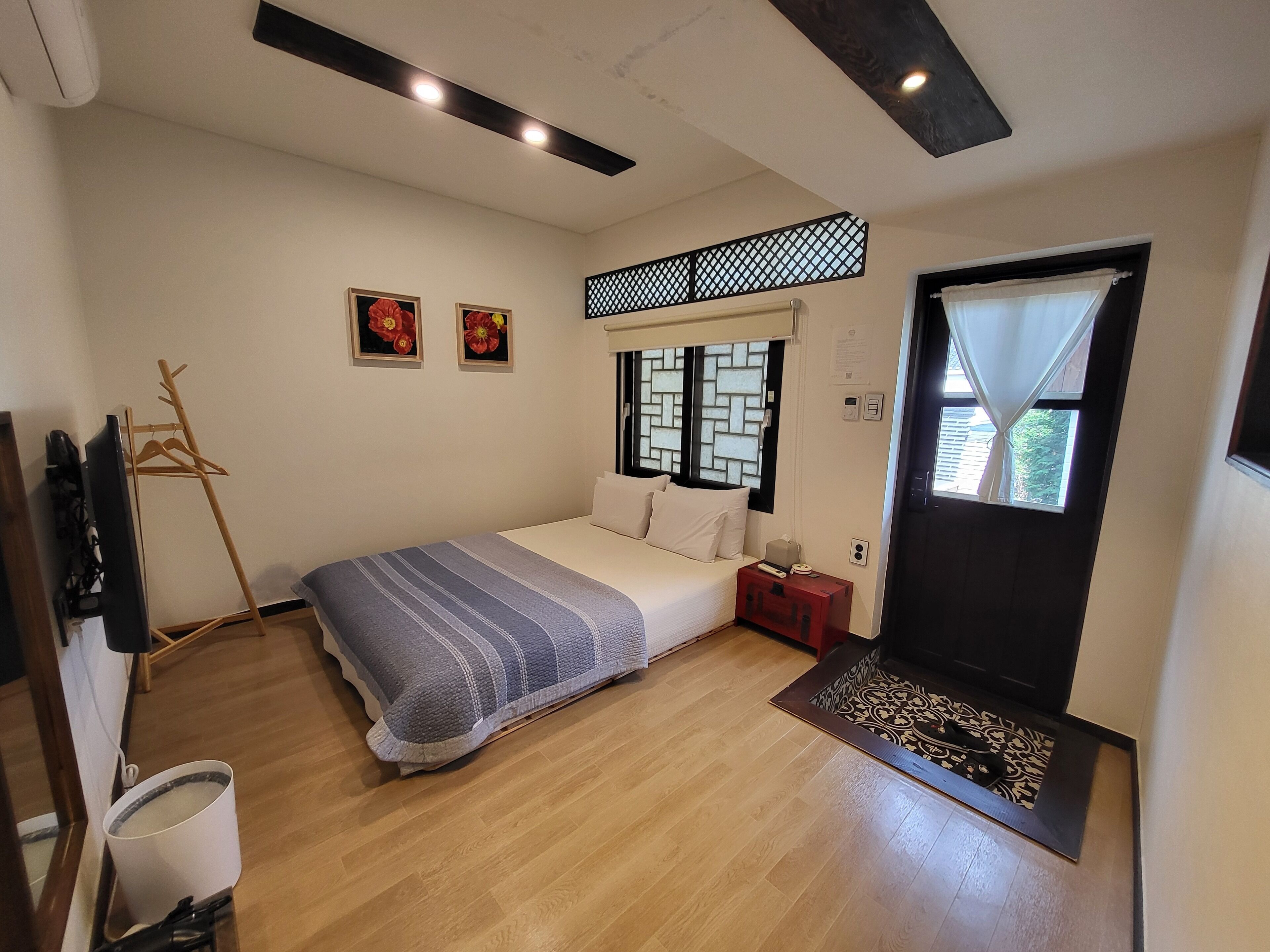 Un dormitorio con cama, suelo de madera, ventana con cortina, mesita de noche y televisor fijado a la pared.