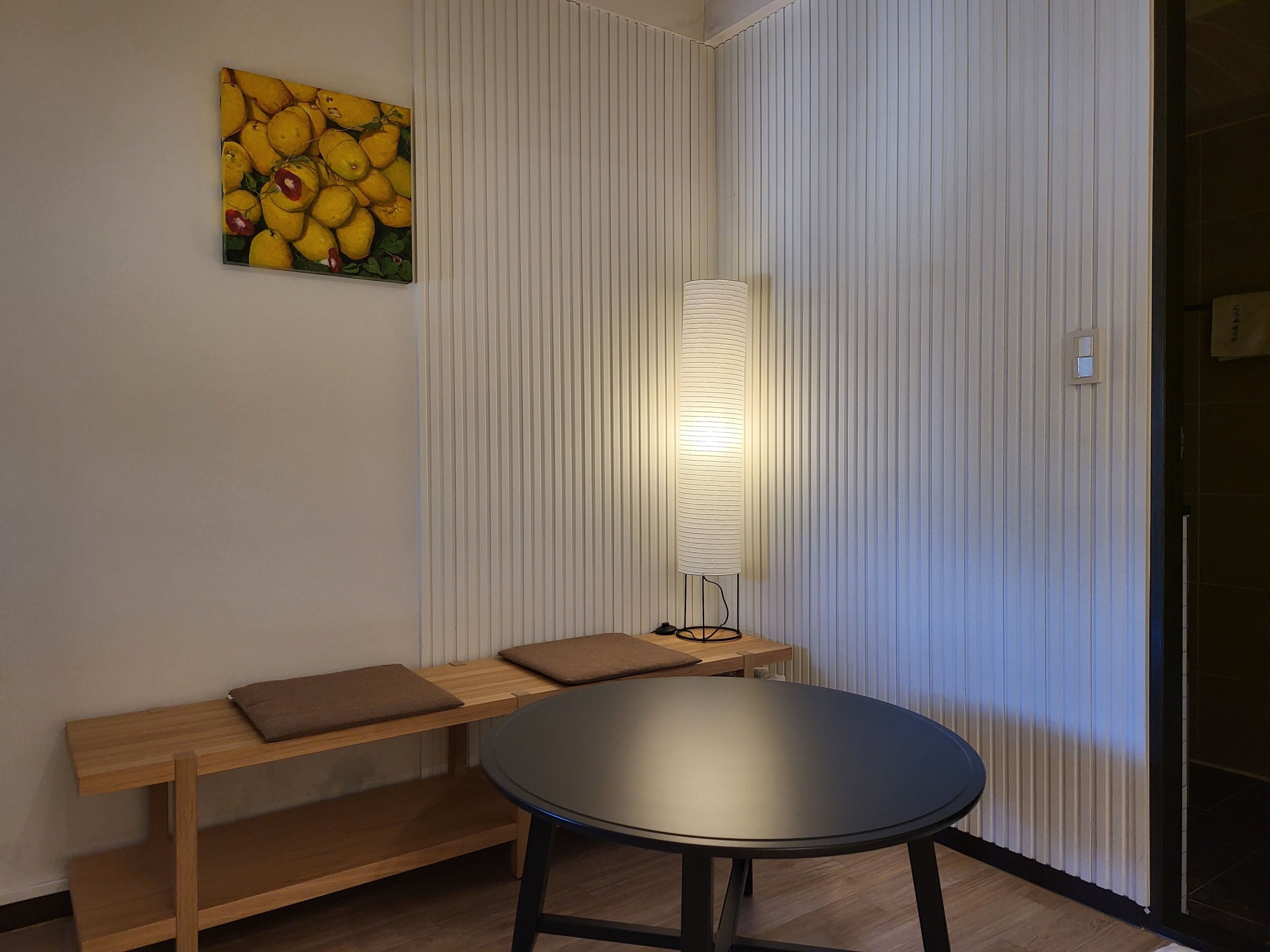 Una habitación con una mesa redonda, un banco con cojines, una lámpara de pared y un cuadro de limones.
