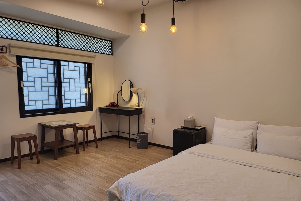 A bedroom with a bed, a desk, a chair, a small table, a mirror, and a nightstand with a lamp.