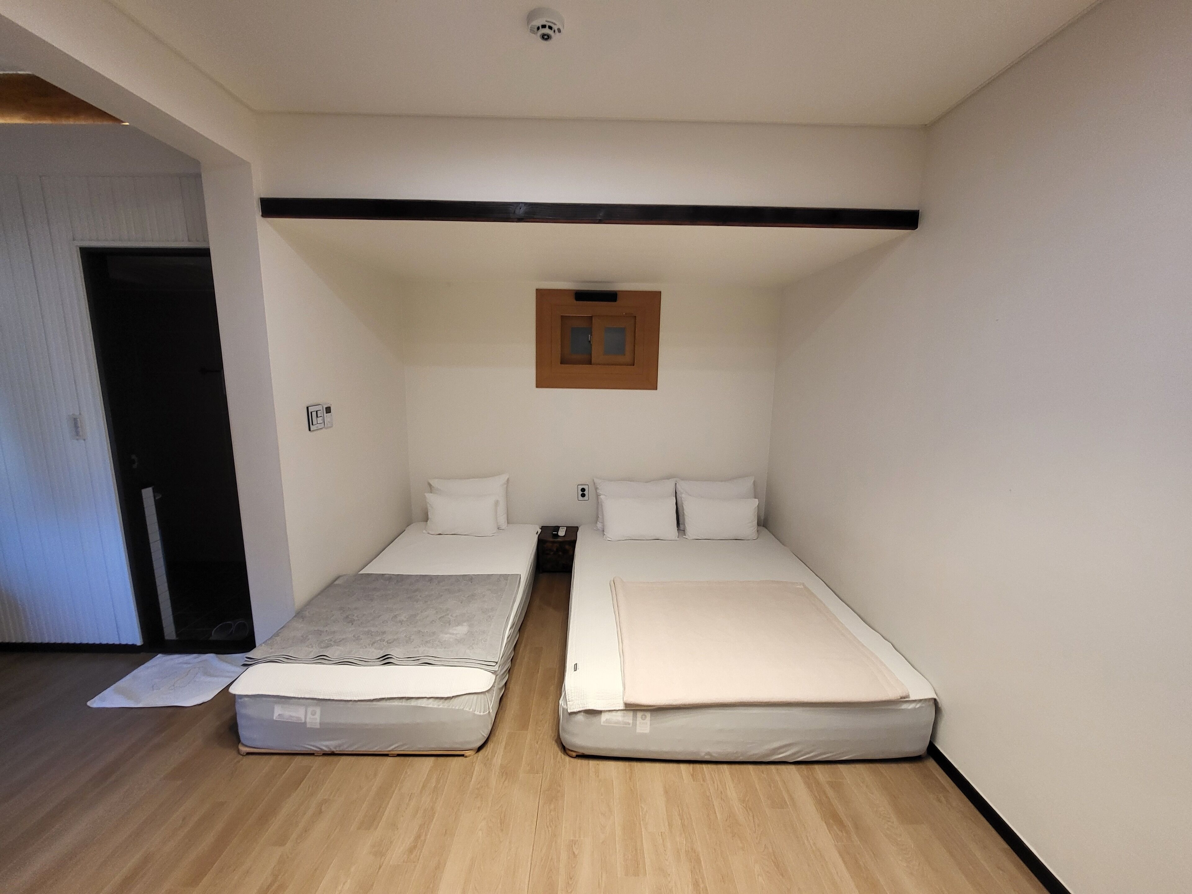 A small, compact room with two single beds, a wooden floor, a ceiling-mounted light, and a door leading to another room.