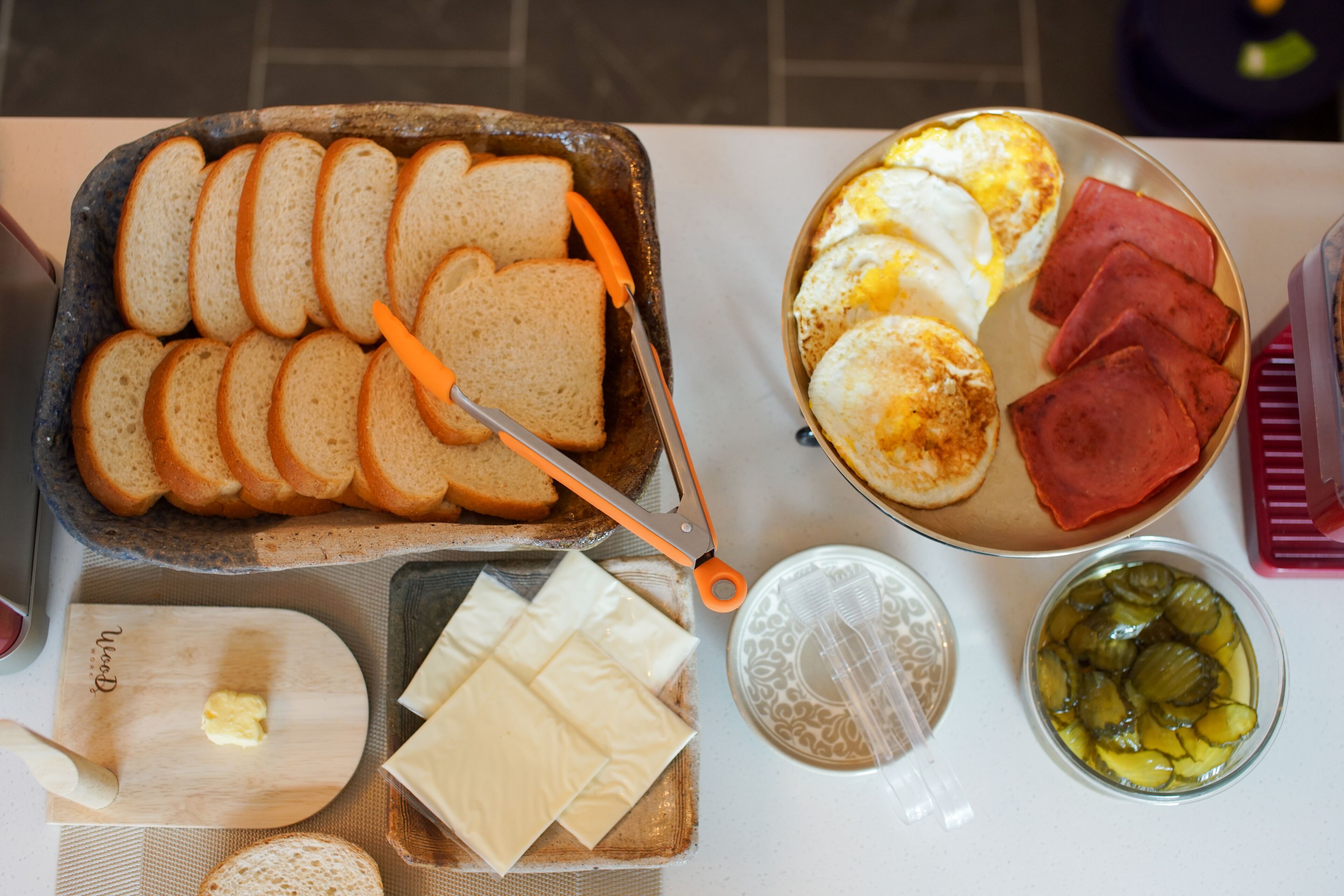 Uma mesa de café da manhã com pão fatiado, queijo, ovos, presunto e picles.