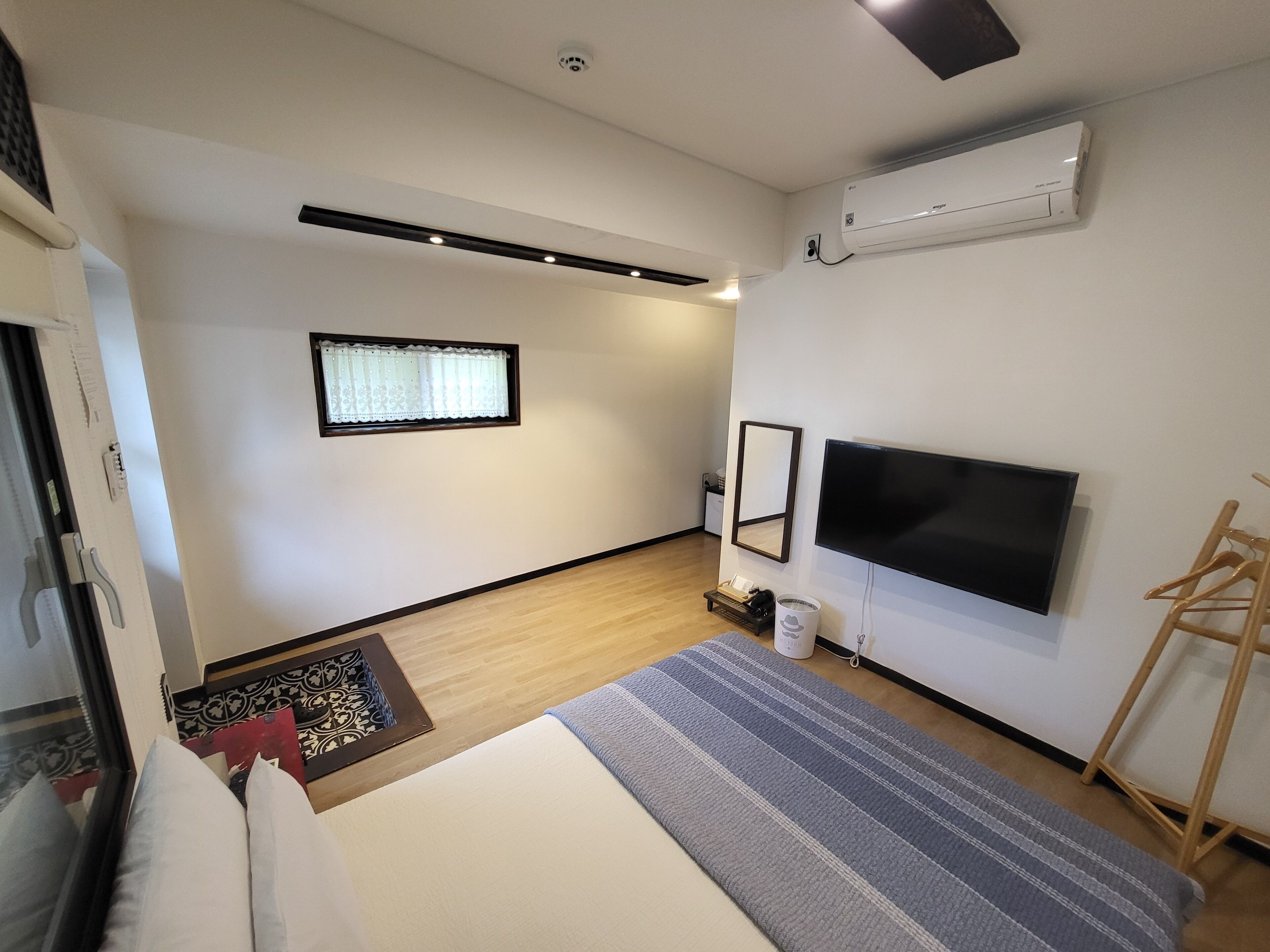 A minimalist hotel room with a bed, a TV, a window with curtains, a wooden floor, and a wall-mounted air conditioner.